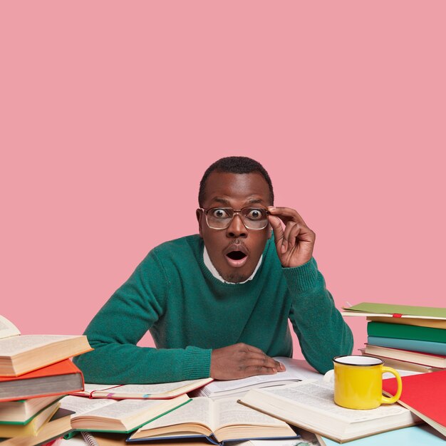 Black young male teacher looks in stupor, surprised to find out about tomorrows lecture, holds hand on rim of glasses