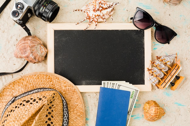 Blackboard near hat with toy boat and money among seashells and camera