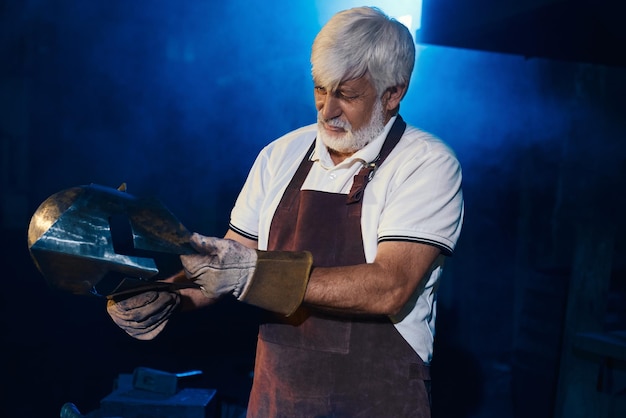 Free photo blacksmith standing at forge and holding gladiator helmet