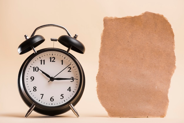 Free photo blank brown torn paper near the black alarm clock against colored backdrop