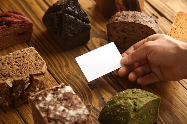 Free photo blank business card of professional artisan baker presented in hand in center of many mixed alternative baked exotic bread samples above wooden rustic table