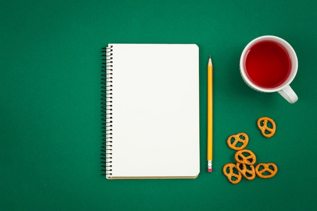 Free photo blank notebook cup of tea and pretzels on green background flat lay