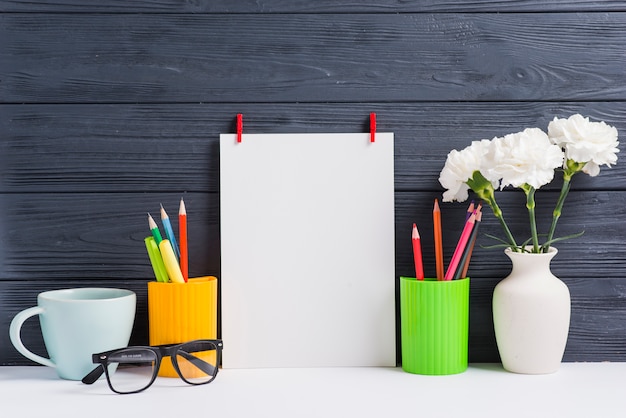 Free photo blank paper; holders; cup; eyeglasses and vase on white desk against wooden background