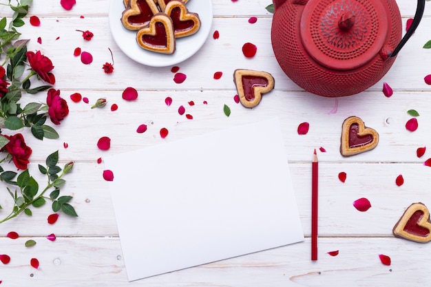 Free photo blank paper with red rose petals on a white surface near a red teapot and heart-shaped cookies