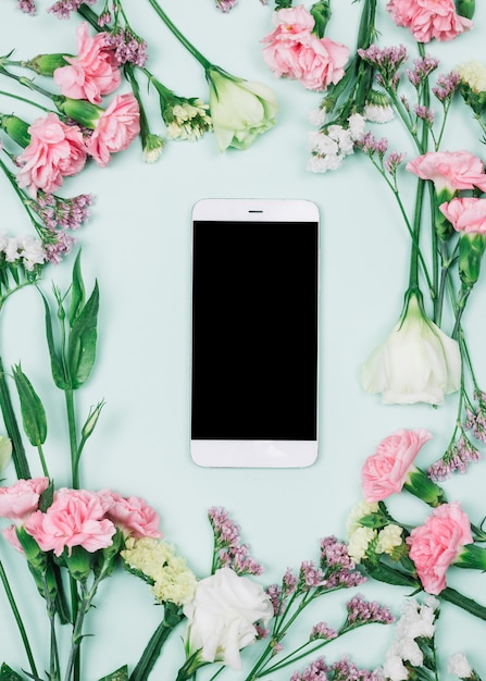 Blank smartphone surrounded with fresh limonium; carnations and eustoma flowers against blue background