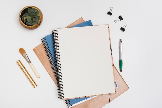 Blank spiral note with make-up brush; mascara; paper clips and pen with potted plant on white desk