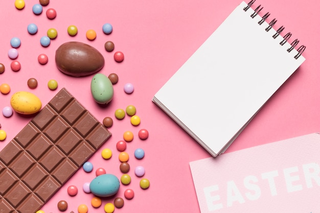 Free photo blank spiral notepad; easter word; easter eggs and multicolored gem candies on pink backdrop