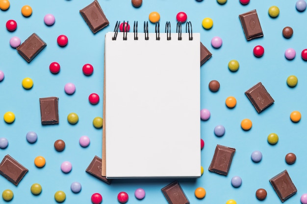 Blank spiral notepad over the gem candies and chocolate pieces on blue background