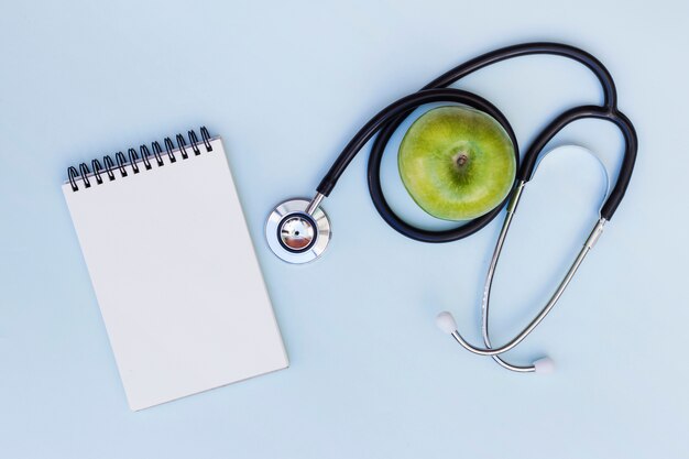 Blank spiral notepad; stethoscope around the green apple on green background