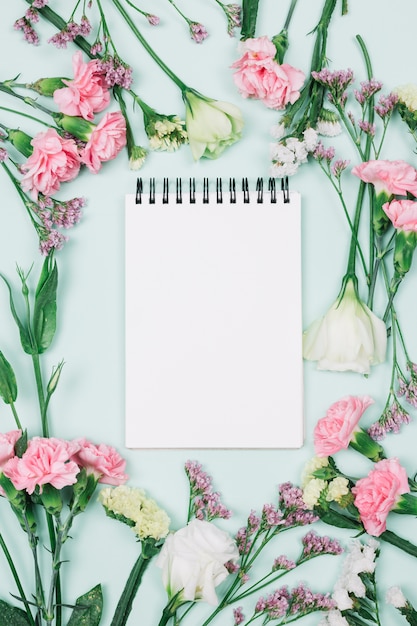 Free photo blank spiral notepad surrounded with limonium; carnations and eustoma flowers against blue background