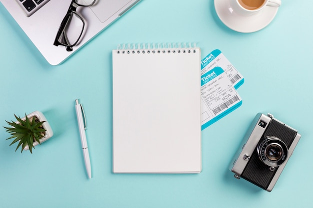 Blank spiral notepad with air tickets surrounded with laptop,eyeglasses,pen,camera,coffee cup on blue desk