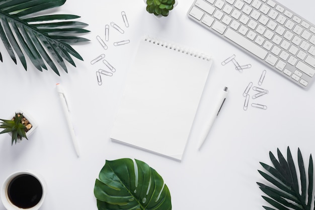 Blank spiral notepad with keyboard; paper clips and green leaves on white background