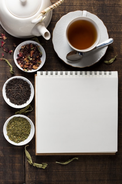 Blank spiral notepad with teapot; tea cup and bowls of herbal tea in white bowl on wooden desk