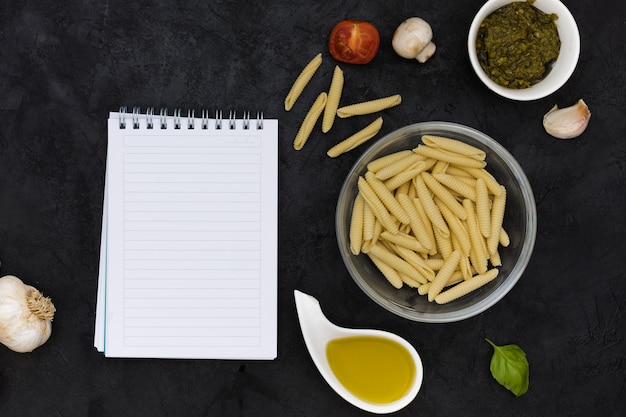 Free photo blank spiral notepad with uncooked garganelli pasta and ingredients on black textured background