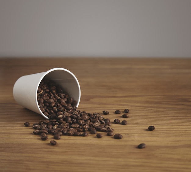 Free Photo blank white paper cup with good roasted coffee beans dropped on thick wooden table in cafe shop