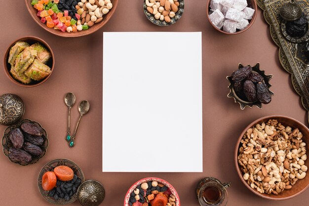 Blank white paper surrounded with circular dried fruits; nuts; lukum; baklava bowl on brown backdrop