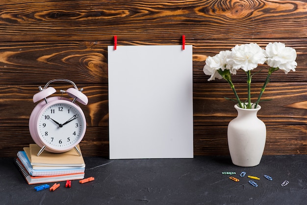 Free photo blank white paper; vase; alarm clock and notebooks against wooden backdrop