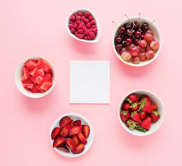 Free photo blank white paper with bowls of red color fruits on pink background