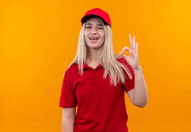 Blinked delivery young woman wearing red t-shirt and cap in dental brace showing okey gesture on isolated orange wall