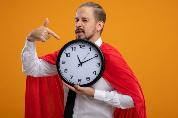 Blinked young superhero guy wearing tie holding and points at wall clock isolated on orange