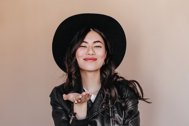 Blissful asian woman in hat gesturing on beige background. Studio shot of romantic curly japanese woman in leather jacket.