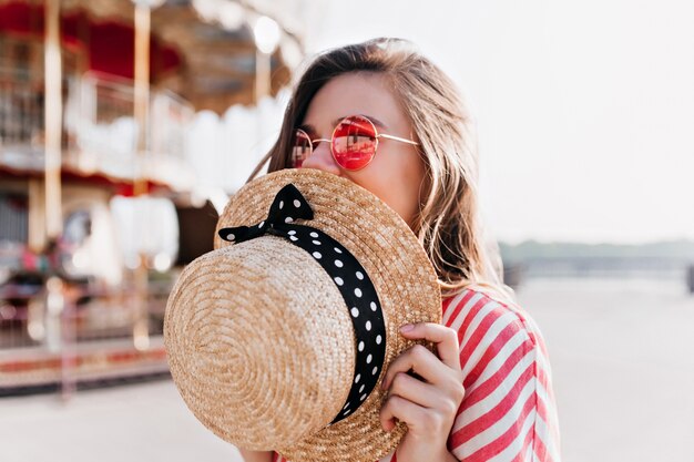 Blissful blonde girl covering face with straw hat while posing in summer day. Outdoor photo of happy young woman in pink sunglasses resting in amusement park.