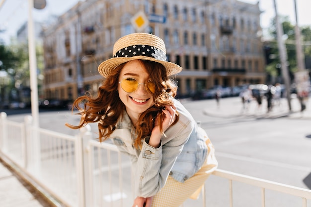 Blissful curly girl in cute hat chilling on the street. Outdoor photo of pretty female model with red hair laughing in summer day.