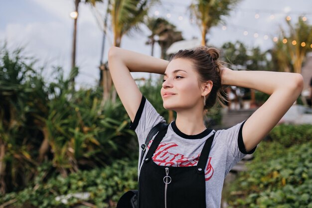 Blissful european girl looking away with romantic smile. Outdoor photo of dreamy young woman in trendy clothes posing with hands up on nature.