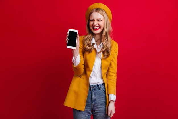 Free Photo blissful french woman holding smartphone with blank screen. front view of elegant blonde girl in beret isolated on red wall.