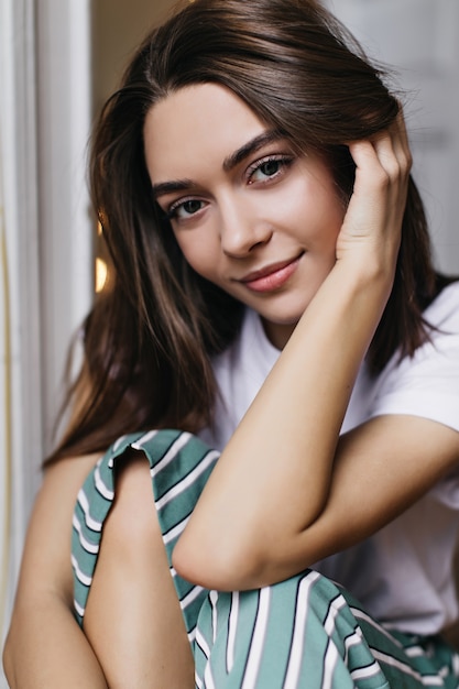 Blissful girl with dark eyes playing with her brown hair. Indoor portrait of pleasant female model with lightly-tanned skin.