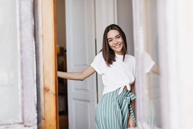 Blissful lady with brown hair smiling while posing at home. Indoor photo of refined woman with trendy hairstyle.