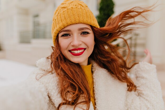 Blissful long-haired woman with red lips making selfie in winter weekend. Blithesome ginger girl in hat expressing happiness.