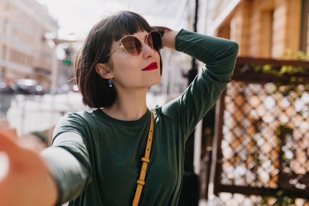 Blissful white woman with short hair making selfie in good spring day. Outdoor photo of interested girl in stylish sunglasses and green sweater.