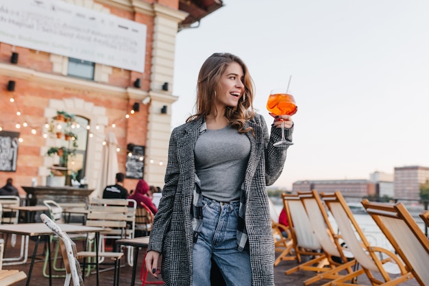 Blissful woman in casual attire raising glass with orange cocktail on city background