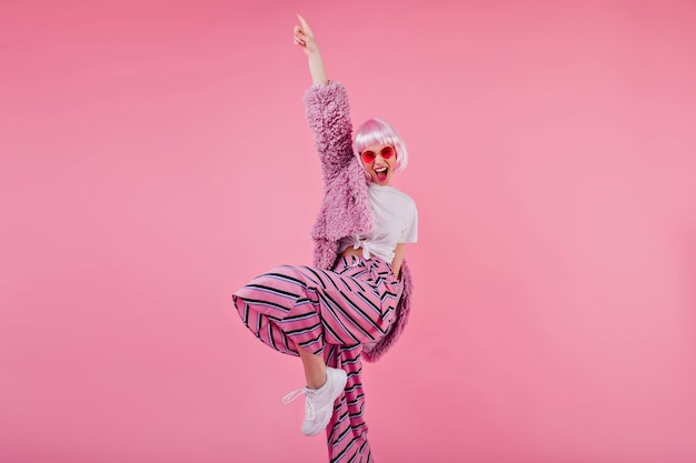 Free photo blissful woman wears striped pants and pink periwig laughing during photoshoot. confident young lady in sunglasses and fluffy jacket funny dancing