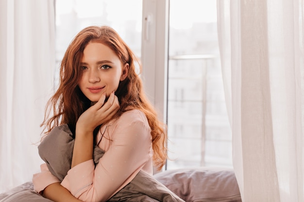 Blithesome caucasian girl in pajama posing in morning. Lovable ginger young lady sitting on bed.