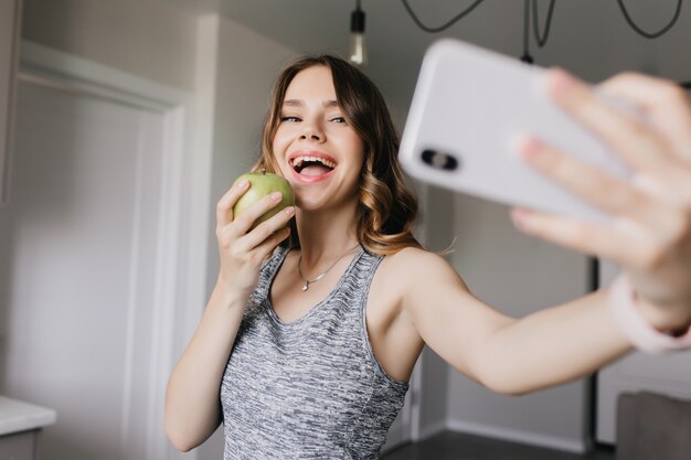 Blithesome caucasian girl taking picture of herself with apple. Laughing lovable woman using smartphone for selfie.