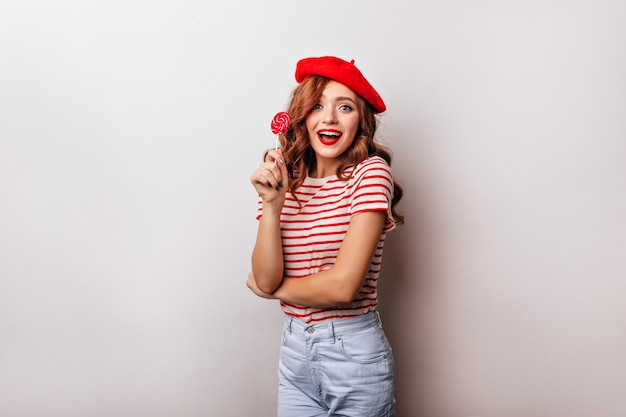 Blithesome curly woman eating lollipop on white wall. Charming french girl in beret posing with candy.