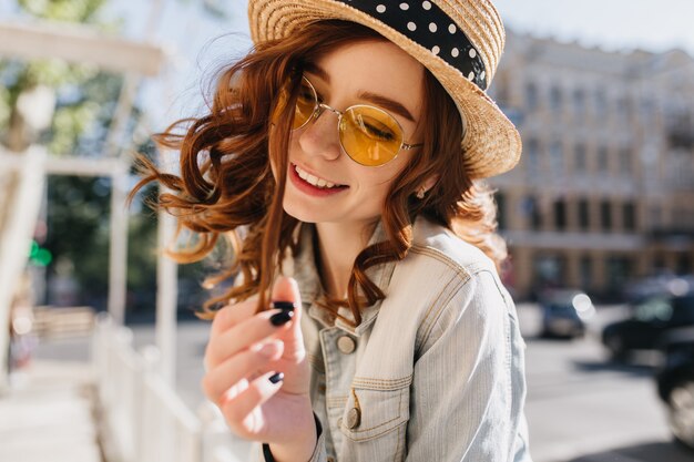 Blithesome european girl playing with her ginger hair with smile. Outdoor shot of happy red-haired lady in summer hat posing on street.