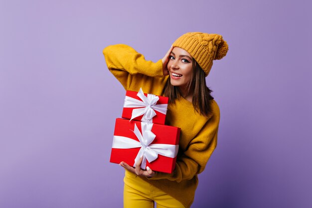 Blithesome girl in yellow hat preparing for christmas. Indoor portrait of glad caucasian lady posing on purple with new year presents.