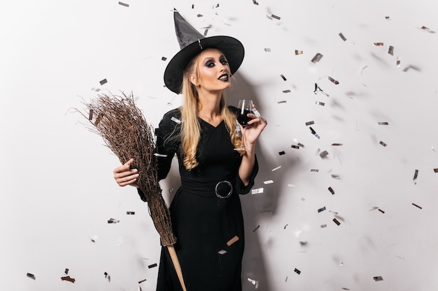 Free Photo blithesome young witch drinking wine. carefree woman in halloween attire posing at party.