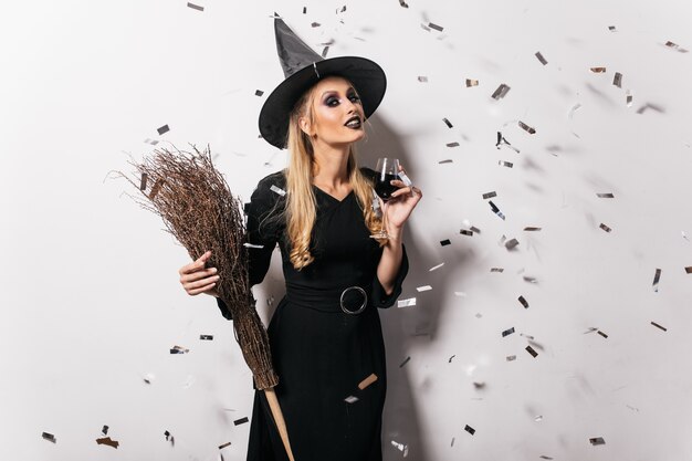 Blithesome young witch drinking wine. Carefree woman in halloween attire posing at party.