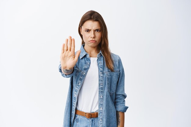 Block Serious woman saying no frowning and extending hand for stop gesture prohibit something disappointing standing against white background