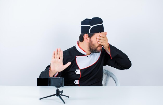 Blogger chef is covering his eyes and showing stop getsure by sitting in front of his mini camera on white background