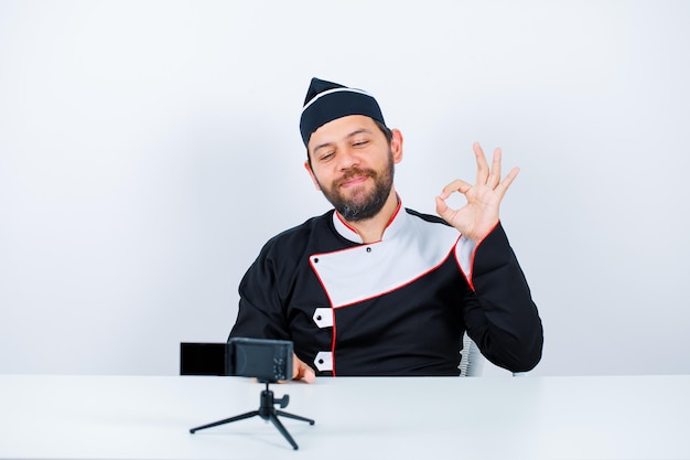 Blogger chef is shosing okay gesture by sitting in front of his mini camera on white background