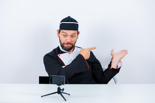 Blogger chef is showing back with ahnd by sitting in front of his mini camera on white background