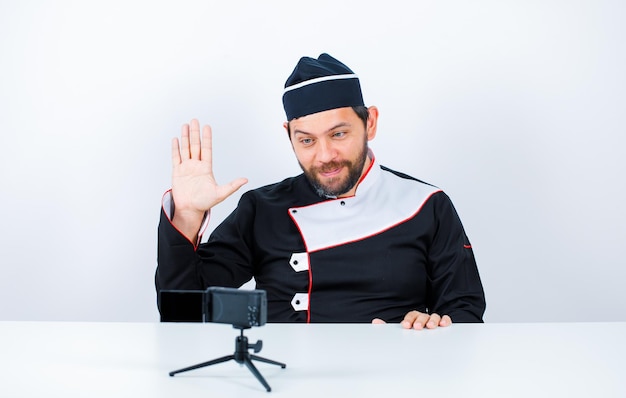 Blogger chef is showing hi gesture by sitting in front of his mini camera on white background