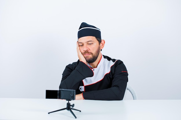 Blogger chef is sitting in front of his mini camera by putting hand on cheek on white background