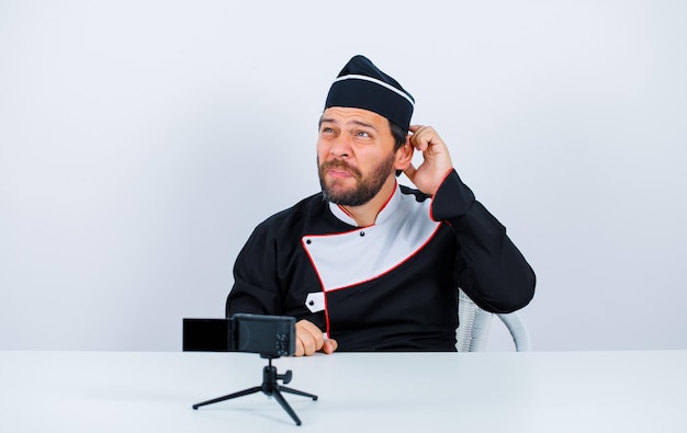 Blogger chef is thinking by holding hand behind his head and sitting in front of his mini camera on white background