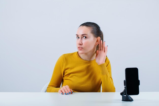 Blogger girl is holding hand behind ear by sitting in front of mobile camera on white background